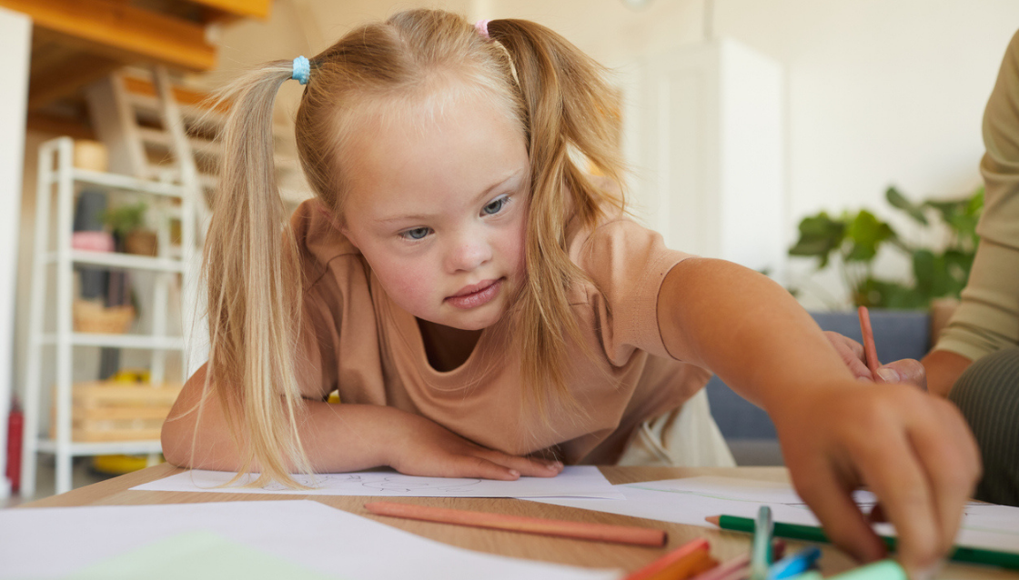 Image of young girl with disability reaching over a desk to grab some pencils and drawing utensils.