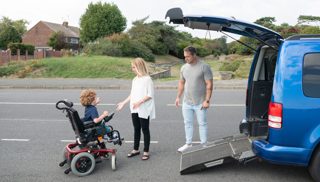 Image of young boy in electric wheelchair and his parents beside an accessible vehicle with a wheelchair ramp extended out of the boot onto the ground.